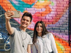 a man and a woman taknig a selfie with his arm extended holding the camera. they are smiling in front of a beautiful and brilliantly colored brick wall mural