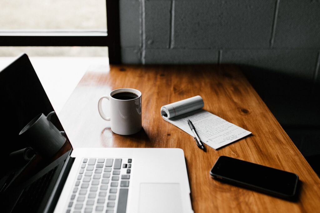 a silver macbook on a wooden table and there is a coffee cup and notepad withe pen and iphone on the table
